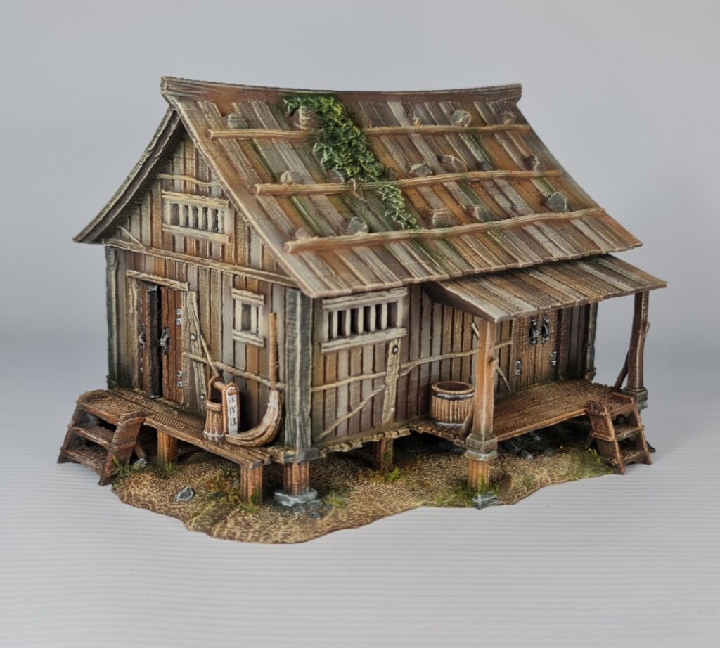 A 28mm scale 3D printed model of an Ishioki-Minka, a traditional Japanese mountain farmhouse. The structure features a unique roof made of flat wooden planks weighted down by numerous realistic stones, a raised timber porch (Engawa) with steps, and weathered wood-grain siding, all set on a stone-pillared foundation.
