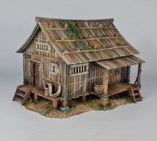 A 28mm scale 3D printed model of an Ishioki-Minka, a traditional Japanese mountain farmhouse. The structure features a unique roof made of flat wooden planks weighted down by numerous realistic stones, a raised timber porch (Engawa) with steps, and weathered wood-grain siding, all set on a stone-pillared foundation.