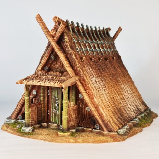 A 28mm scale 3D printed model of a Chigi-Minka, an ancient Japanese dwelling. The structure is characterized by a steep A-frame thatched roof topped with prominent crossed wooden finials (Chigi) and horizontal logs (Katsuogi). It features a sheltered entrance with timber pillars, rustic wooden doors, and a stone-lined foundation, reflecting a high-status or spiritually significant historical residence.