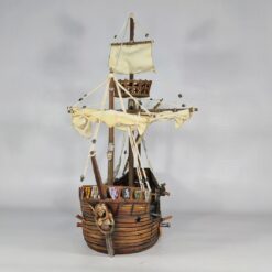 Front-view photograph of a fully assembled and painted 3D printed model of a 15th-century Caravel sailing ship. The model features a detailed wooden plank hull, a mermaid figurehead, rigging, two masts with fabric-like sails, and decorative shields on the bow.