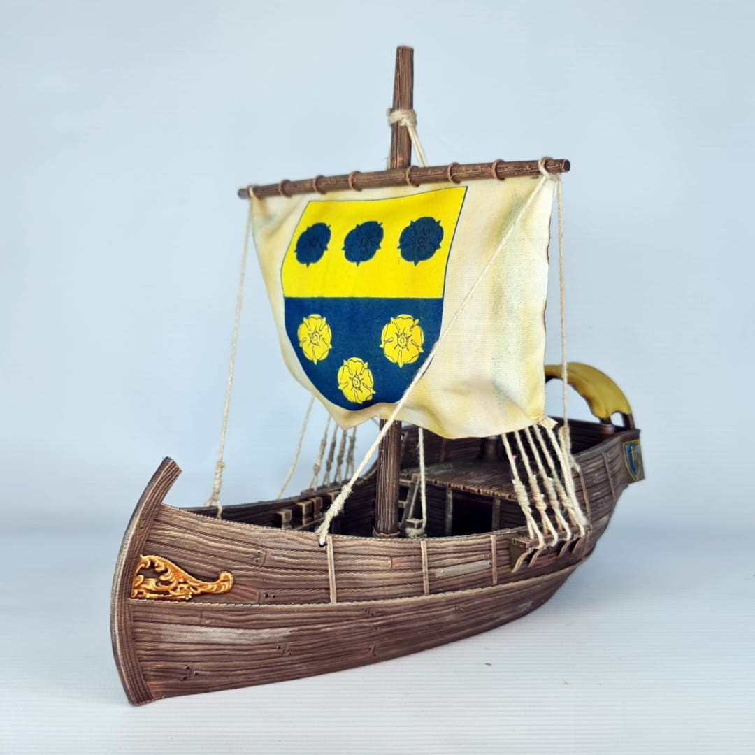 3D printed and painted model of a medieval merchant cog ship, front view. The ship has a detailed wooden hull, a single mast, and a square sail with a blue and yellow coat of arms.