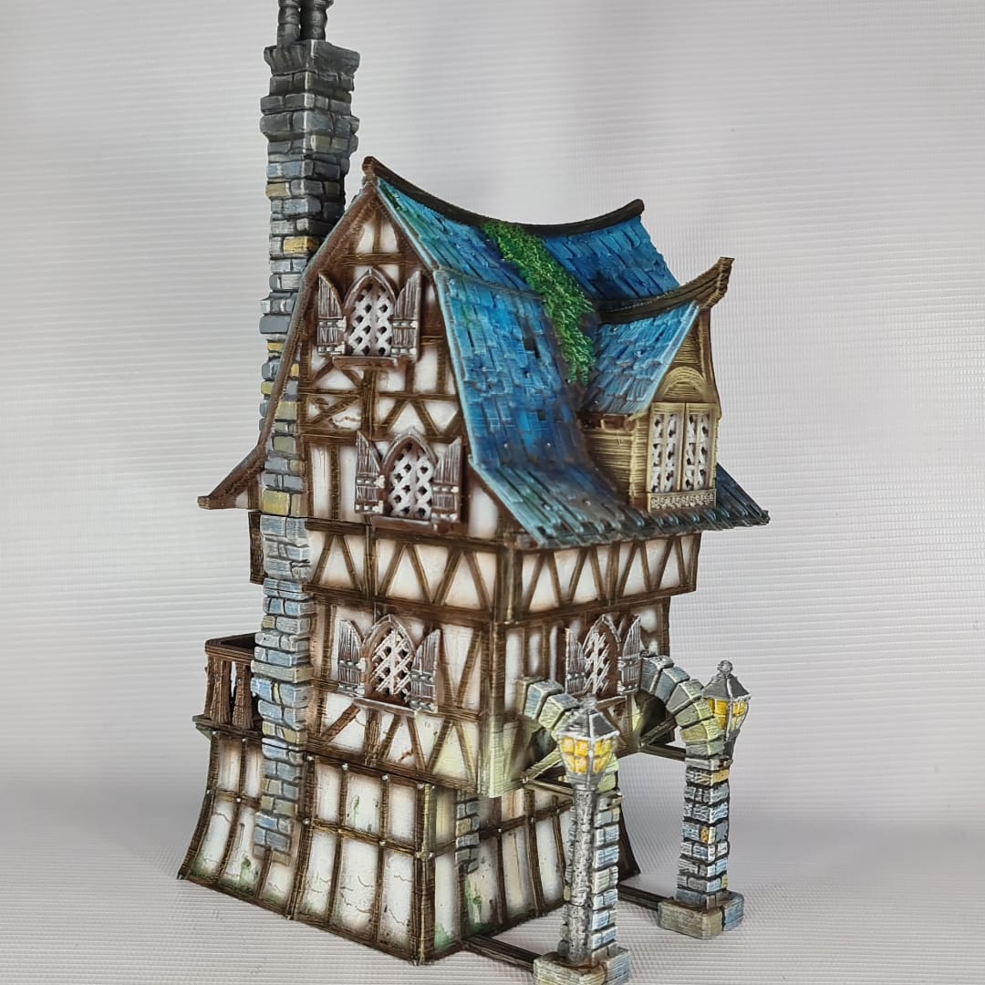 3D printed and painted model of a multi-story medieval fantasy house featuring half-timbered walls, a steep blue roof, stone pillars with lamps, and an external wooden staircase.