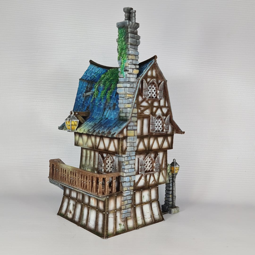 3D printed and painted model of a multi-story medieval fantasy house featuring half-timbered walls, a steep blue roof, stone pillars with lamps, and an external wooden staircase.