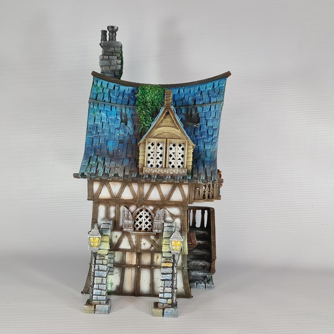 3D printed and painted model of a multi-story medieval fantasy house featuring half-timbered walls, a steep blue roof, stone pillars with lamps, and an external wooden staircase.
